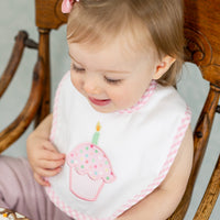 Child wearing a bib with a pink cupcake design sitting in a wooden highchair, 3 Marthas