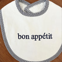 White bib with navy text 'bon appetit' and checkered trim on a wooden table