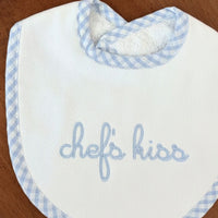Baby bib with blue checkered trim and 'Chef's Kiss' text on a wooden table.