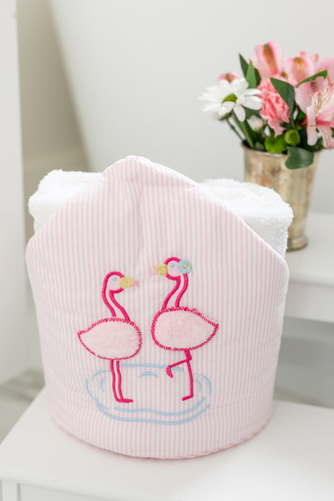 Pink hooded towel with flamingo design on a white surface with flowers in the background, 3 Marthas