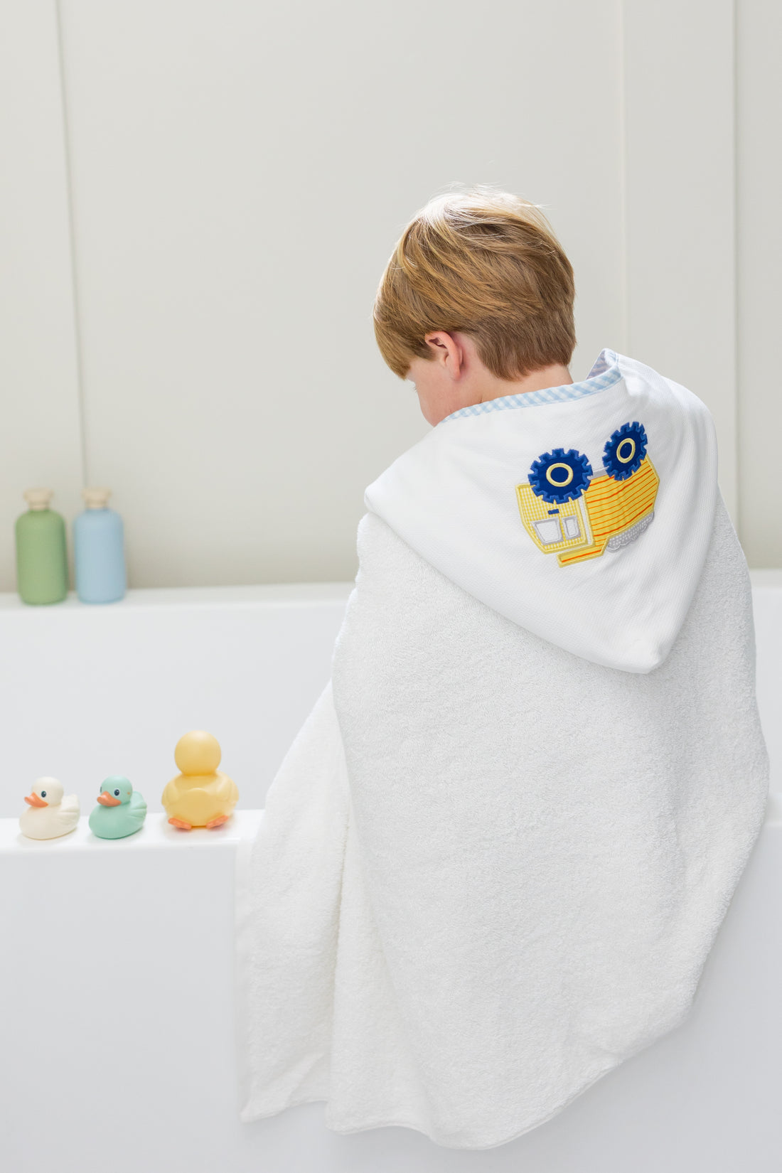 Child wrapped in a white hooded towel with a yellow dump truck, sitting on a bathtub with colorful rubber ducks, 3 Marthas
