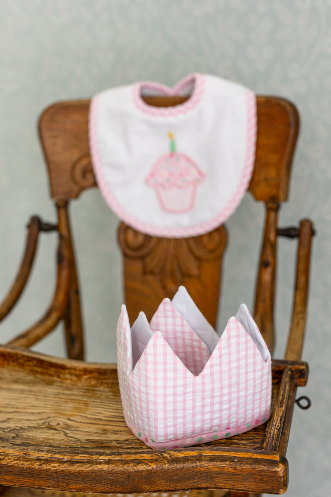 Pink bib with cupcake design and matching crown on a wooden chair against a gray background, 3 Marthas