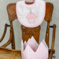 Pink bib with cupcake design and matching crown on a wooden chair against a gray background, 3 Marthas