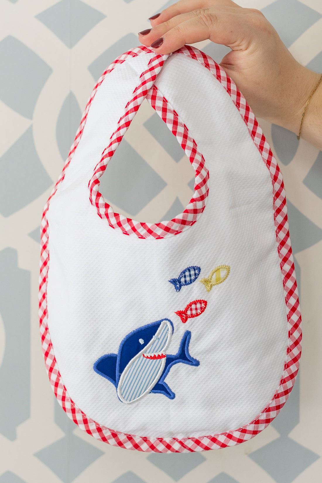 Baby bib with shark design and red checkered trim held by a hand against a patterned background, 3 Marthas