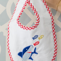 Baby bib with shark design and red checkered trim held by a hand against a patterned background, 3 Marthas