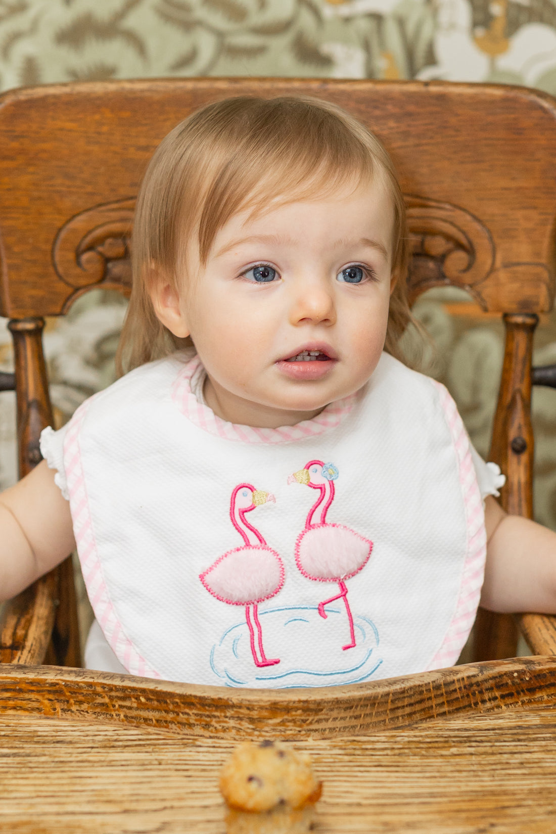 Child wearing a bib with flamingo design sitting in a wooden high chair, 3 Marthas