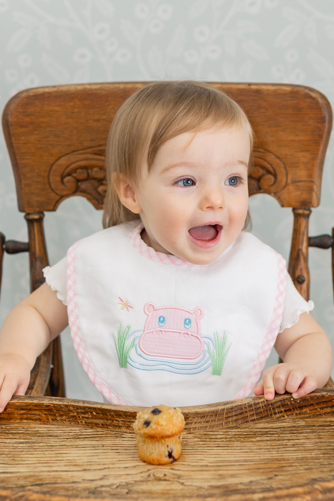 Baby wearing a bib with a pink hippo design, sitting in a wooden high chair, 3 Marthas