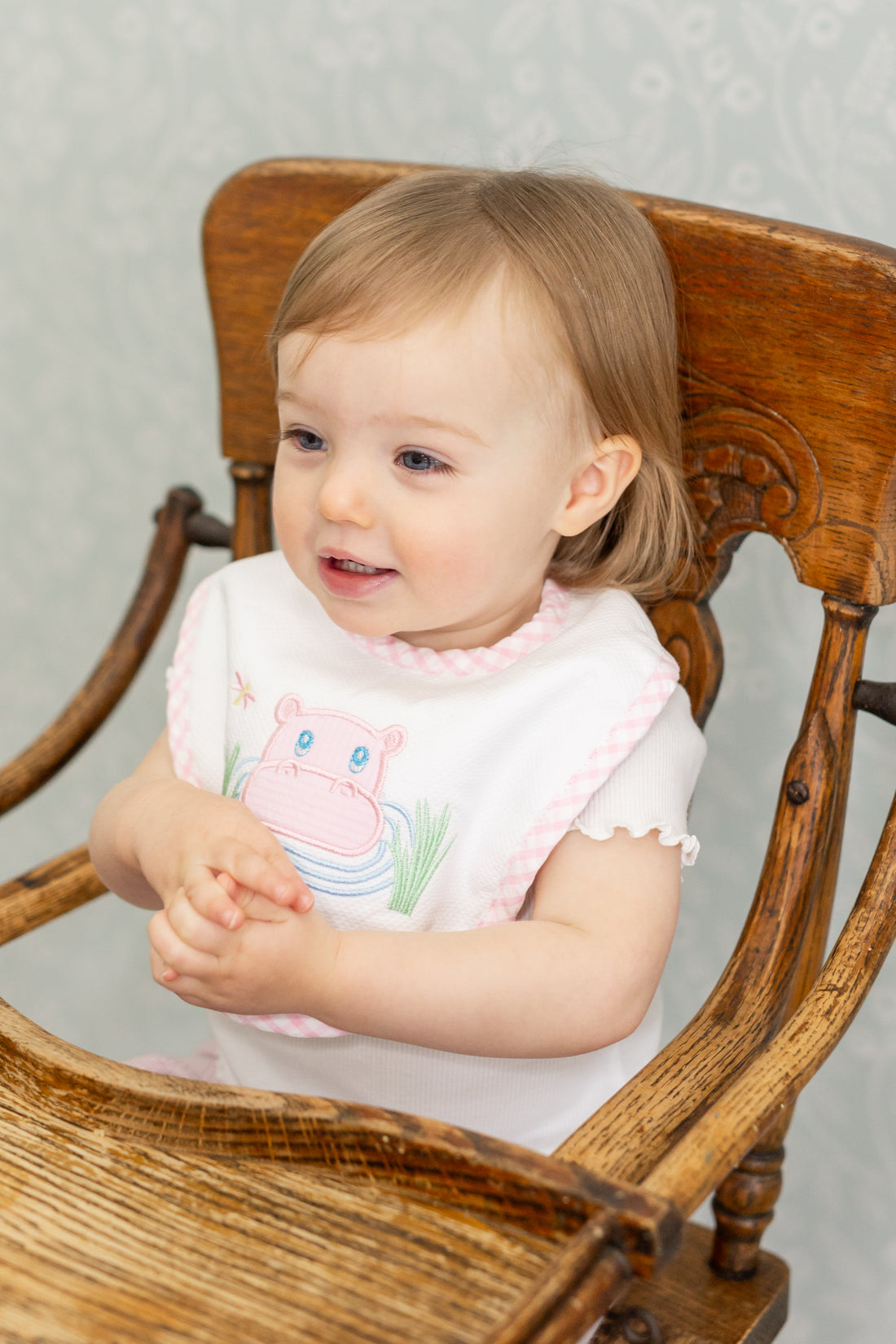 Child wearing a bib with a pink hippo applique in a wooden high chair, 3 Marthas