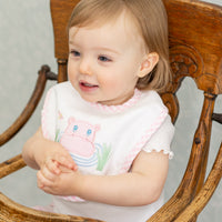 Child wearing a bib with a pink hippo applique in a wooden high chair, 3 Marthas
