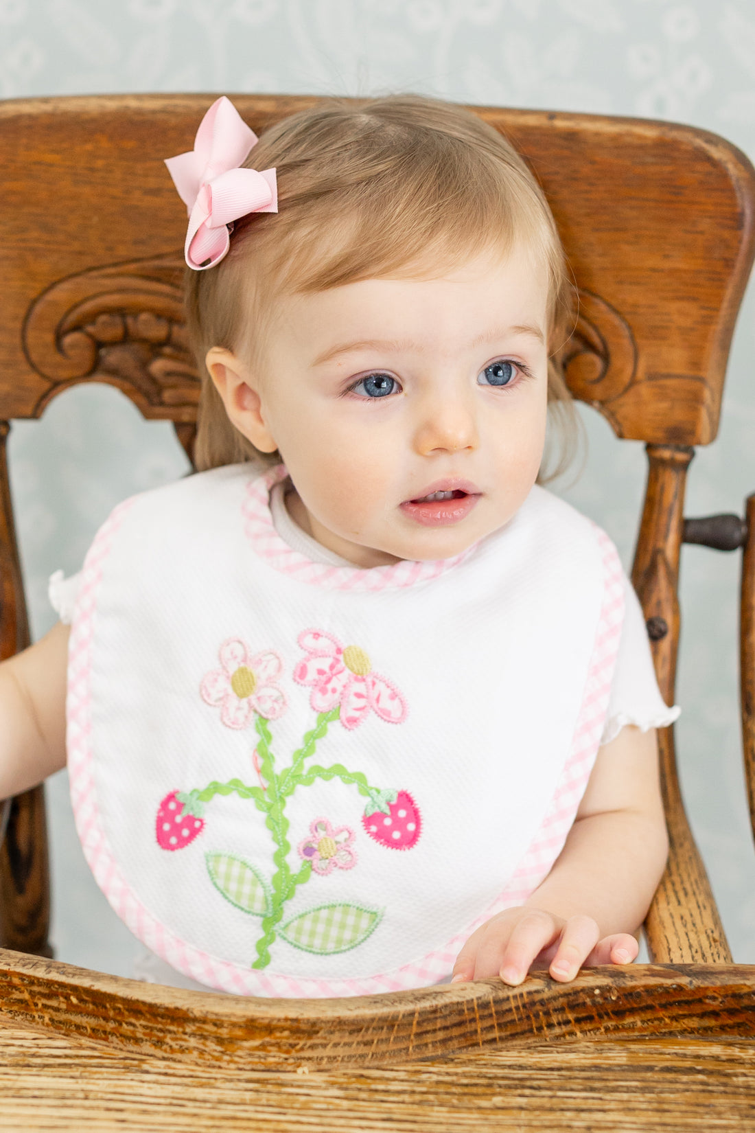 Baby wearing a bib with strawberry floral design sitting in a wooden high chair, 3 Marthas