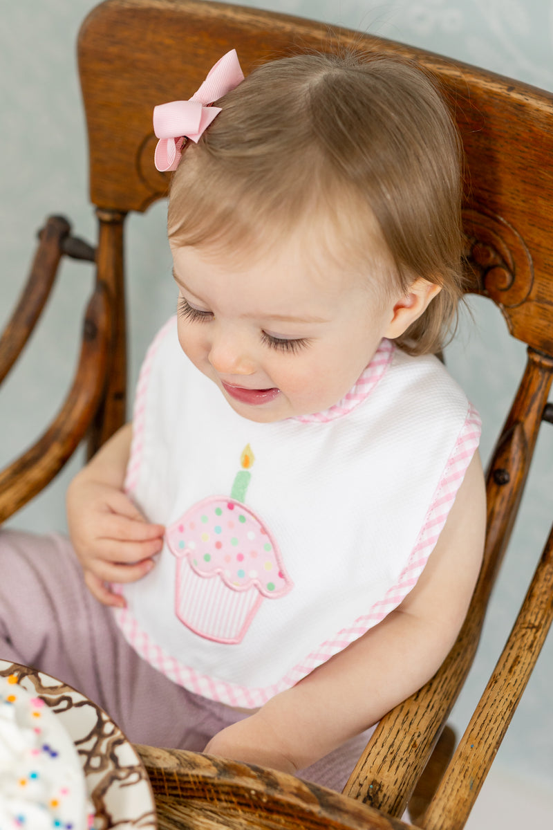 Child wearing a bib with a pink cupcake design sitting in a wooden highchair, 3 Marthas