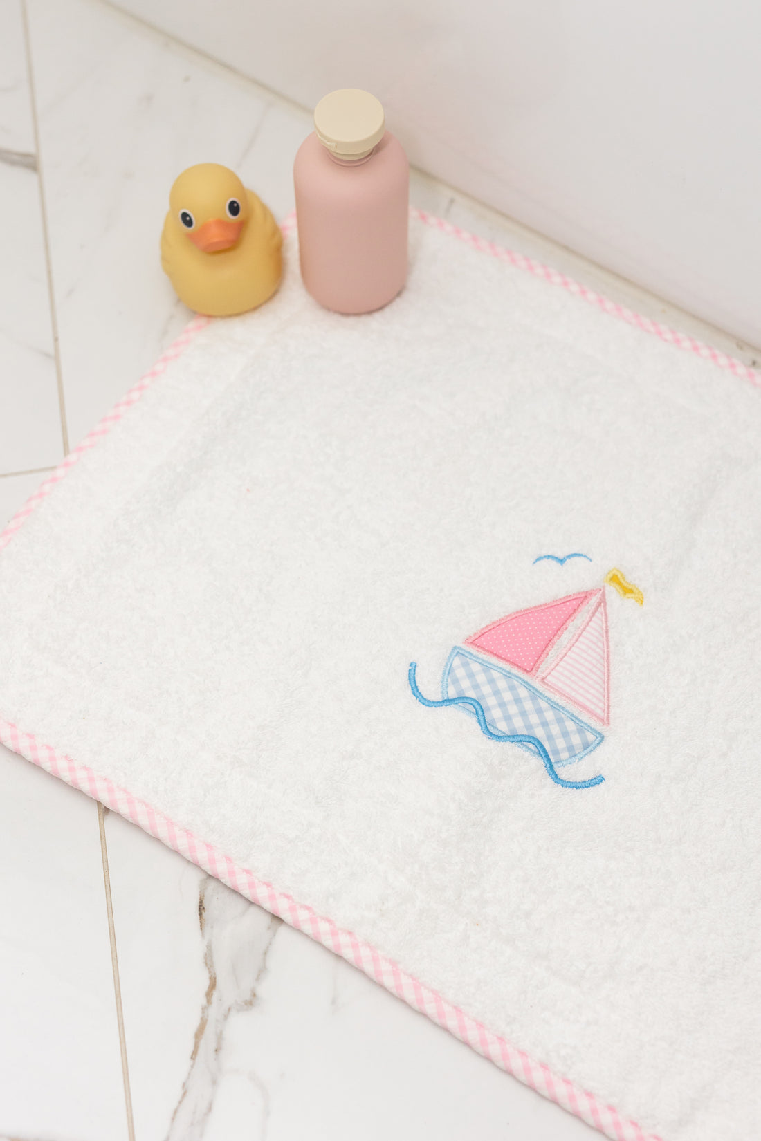 Bath mat with pink sailboat design, rubber duck, and bottle on a tiled floor, 3 Marthas