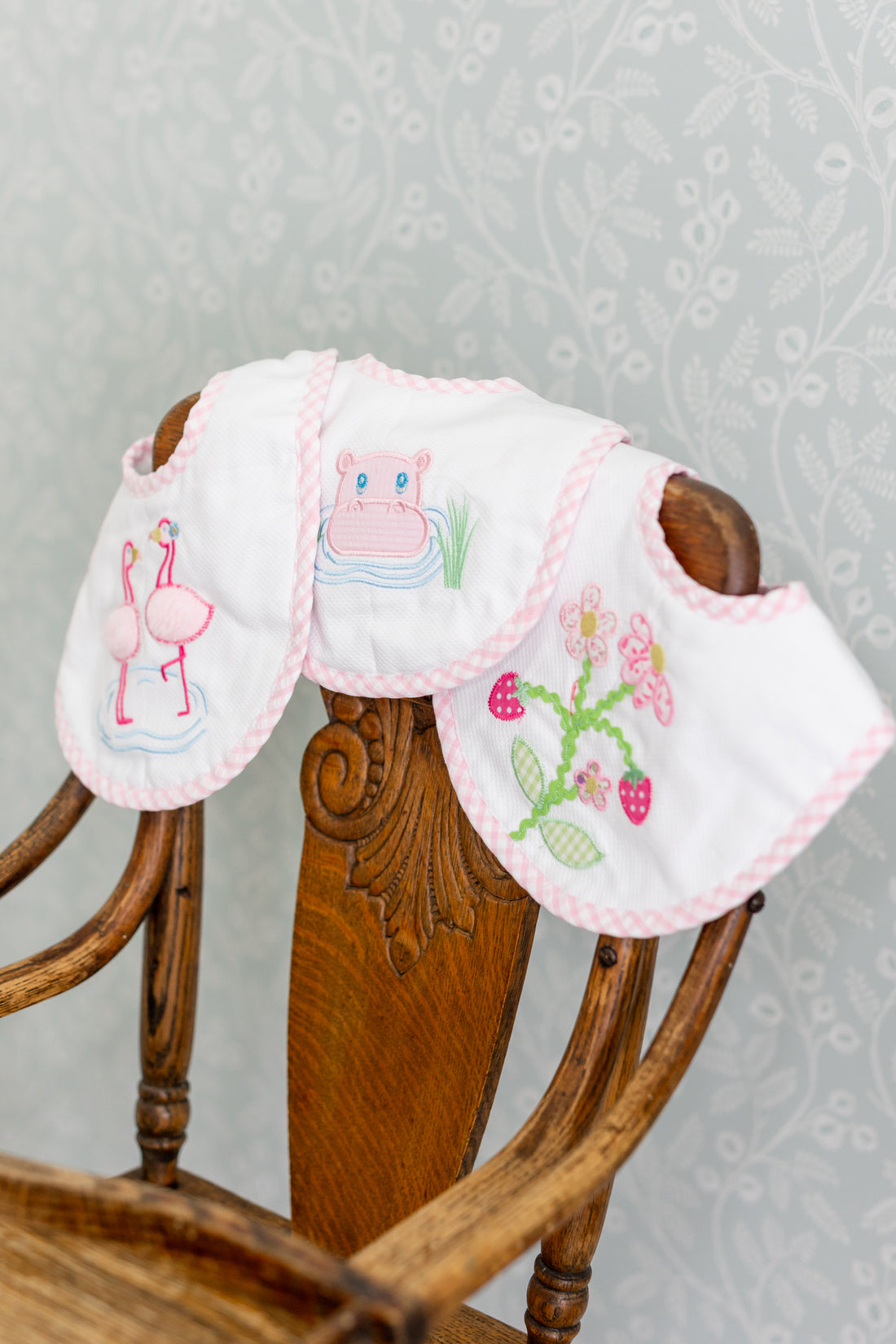 Three baby bibs with embroidered designs on a wooden chair against a floral-patterned wall, 3 Marthas