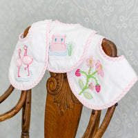 Three baby bibs with embroidered designs on a wooden chair against a floral-patterned wall, 3 Marthas